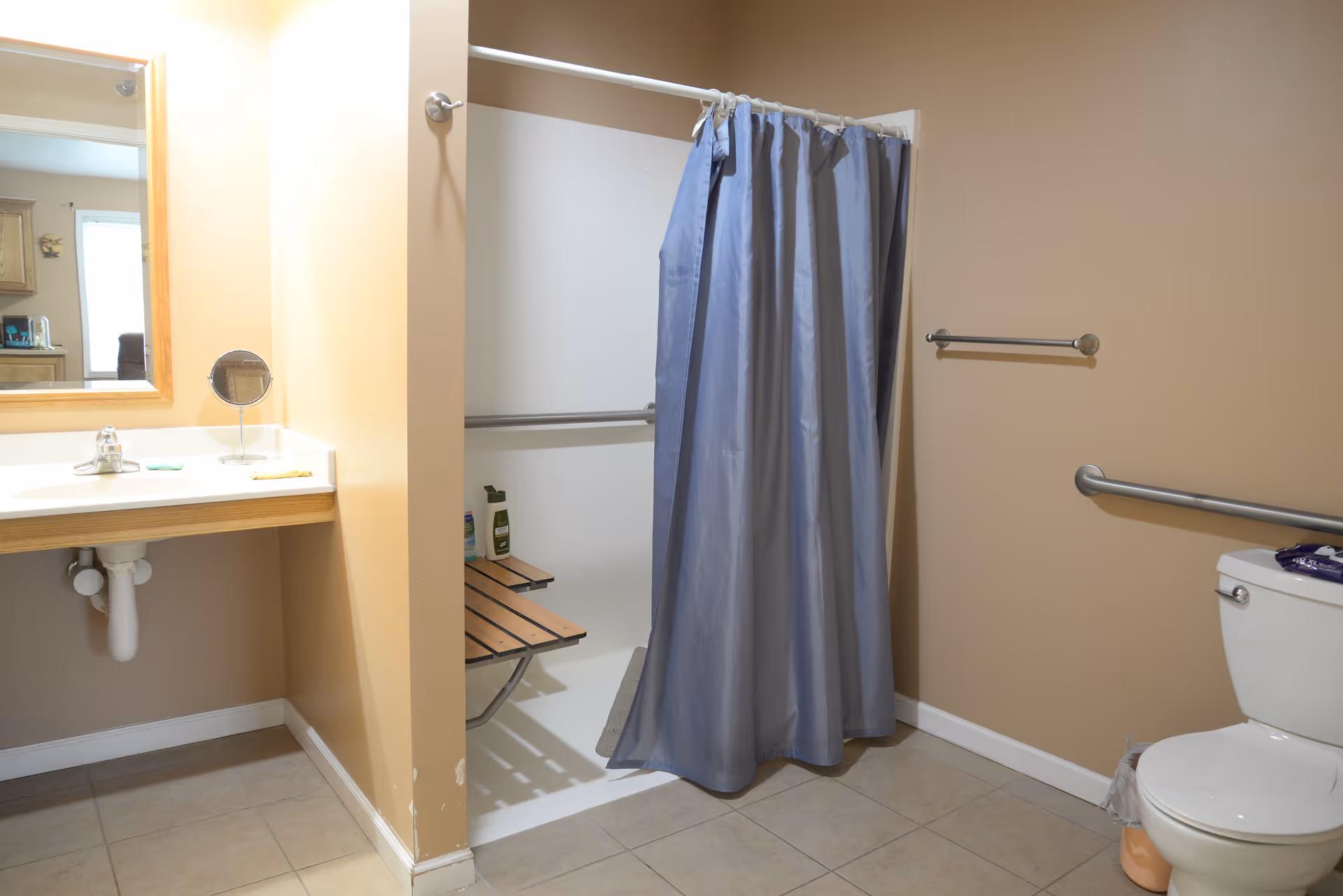 Accessible bathroom with a shower stall and blue curtain, sink and mirror, grab bars, and a toilet.