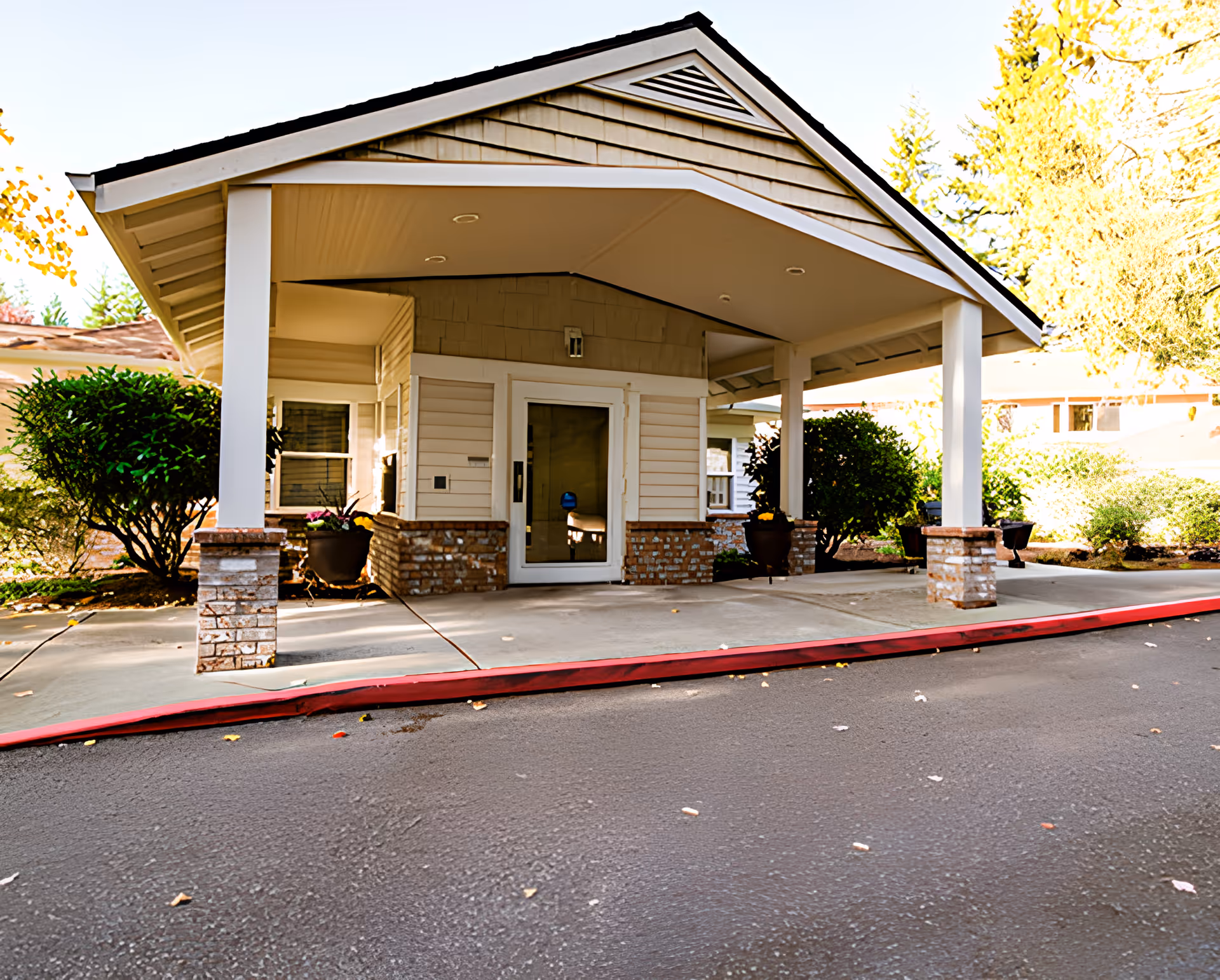 Covered entrance/porte-cochère of a senior living facility with columns, potted plants, and a glass entry door.