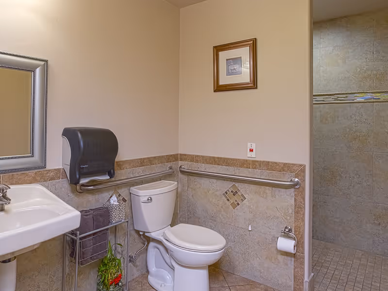 A clean and accessible bathroom with a white toilet, a white sink with a silver faucet, a wall-mounted black paper towel dispenser, and stainless steel grab bars on the walls. The walls are tiled halfway up with beige tiles featuring a decorative diamond pattern. There is a framed picture hanging on the wall above the toilet. To the right, there is an open doorway leading to a tiled shower area.