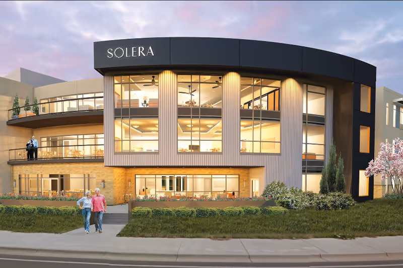 Front exterior of a modern three-story building labeled "SOLERA" with large windows, landscaping, and two people walking on the sidewalk.