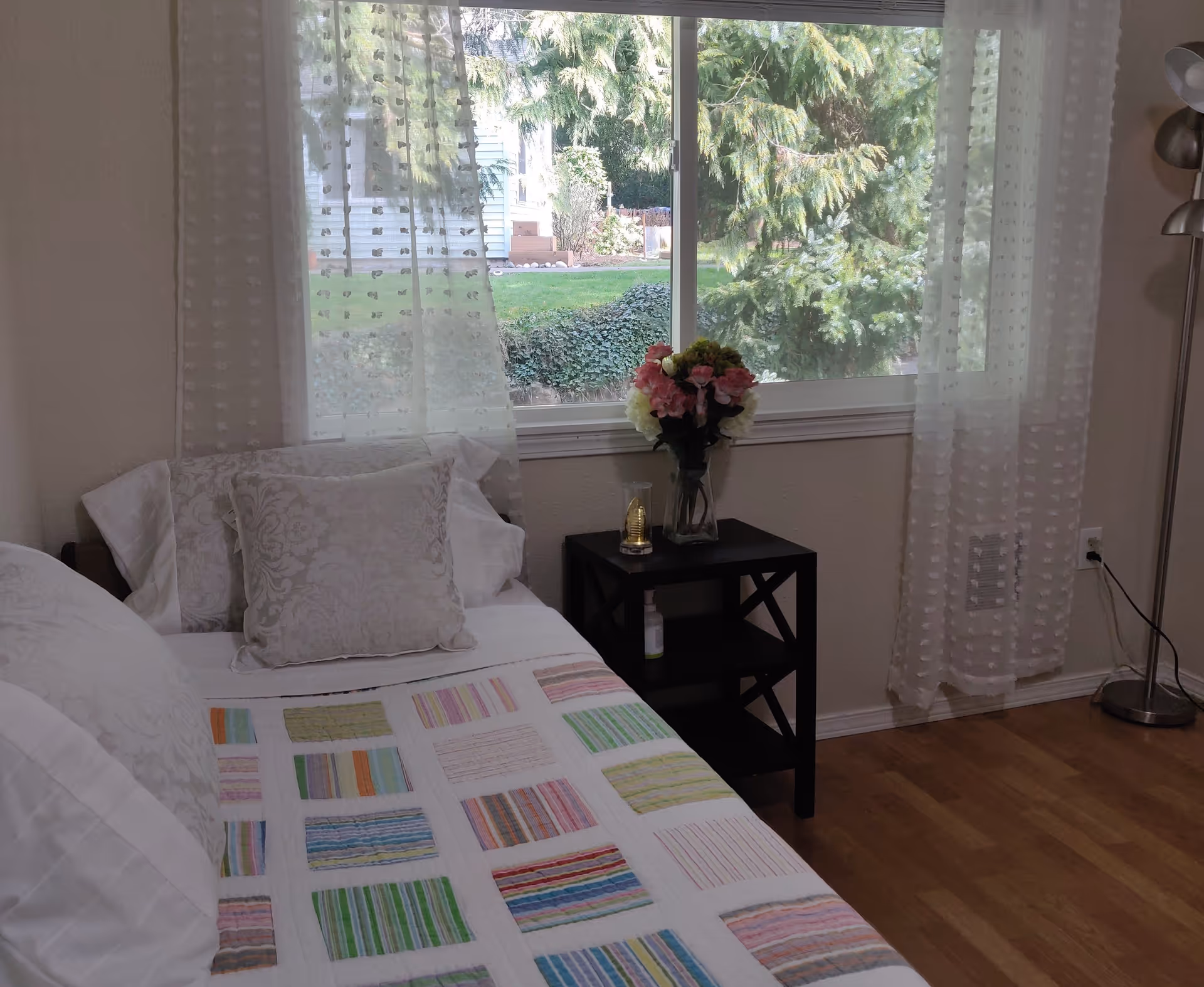 A cozy bedroom with a bed covered in a colorful patchwork quilt and decorative pillows. Next to the bed is a black nightstand with a vase of flowers and a small decorative item. Sheer white curtains frame a large window showing greenery outside. A floor lamp stands in the corner on a wooden floor.