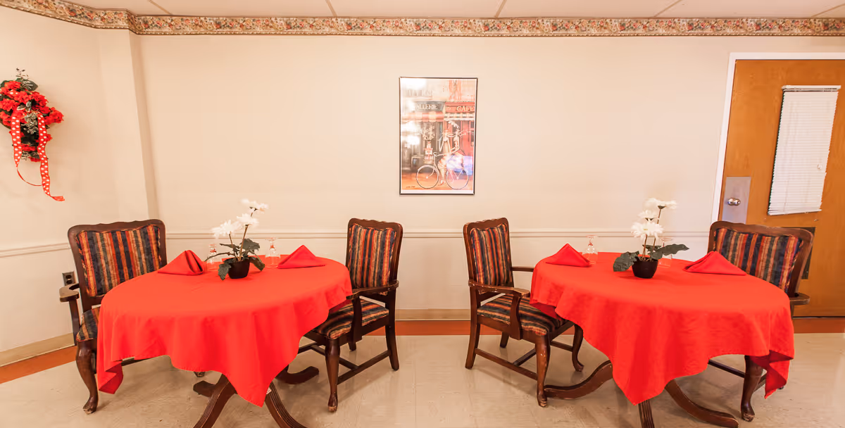 Two round tables covered with red tablecloths, each with a small potted white flower centerpiece and red folded napkins. Four wooden chairs with striped upholstery surround the tables. A framed picture hangs on the beige wall behind the tables, and a wooden door with a small window is visible on the right. A decorative red and green wreath with a red polka dot ribbon is mounted on the left wall.