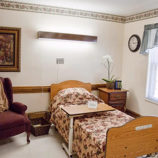 A single bed with floral patterned bedding in a small room. Next to the bed is a wooden nightstand with a potted white orchid and some books. A small overbed table is positioned over the bed with a welcome card on it. To the left of the bed is a maroon upholstered armchair and a woven basket on the floor. The walls are light-colored with a floral border near the ceiling, a framed painting, a clock, and a window with a blue valance.