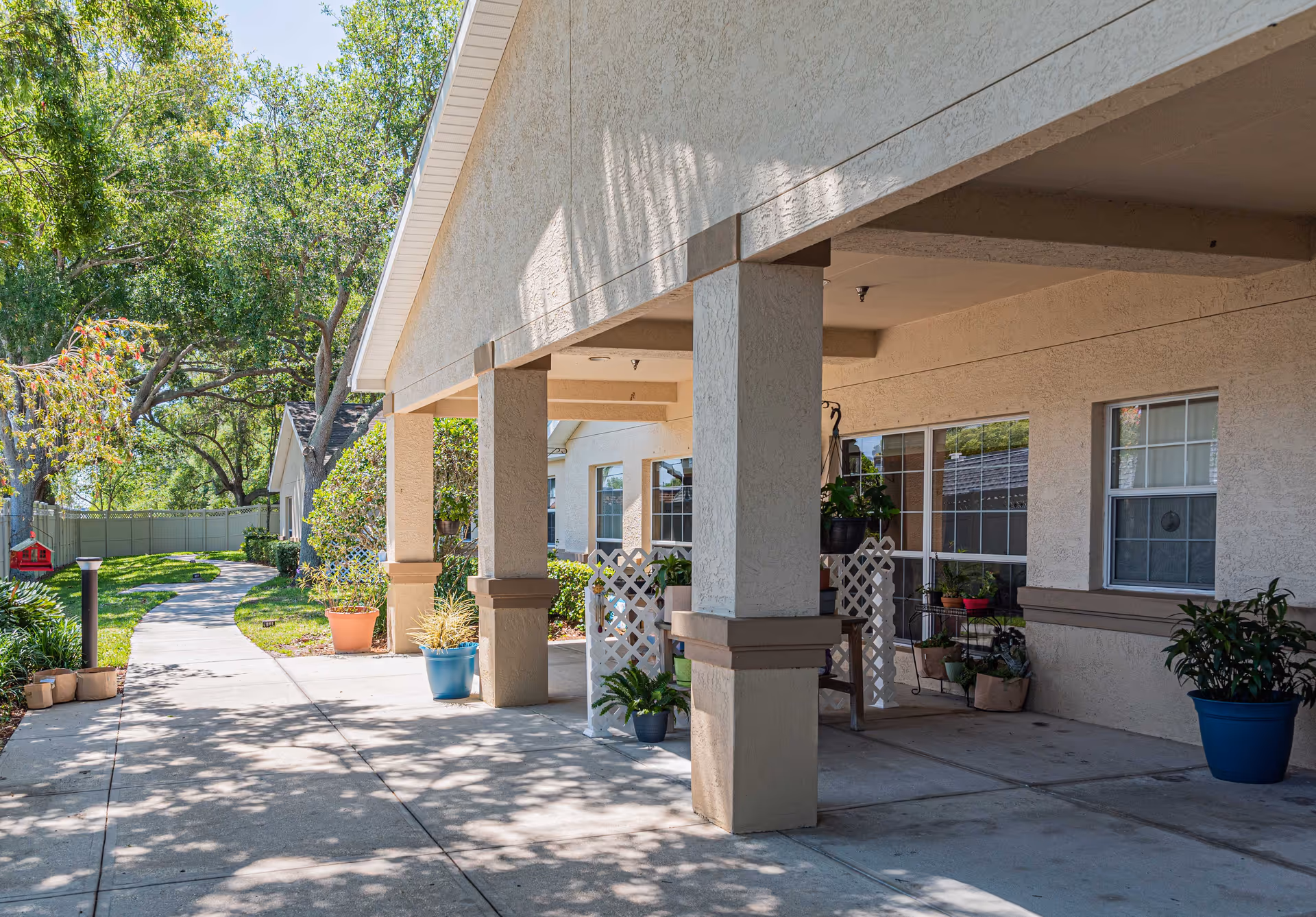 Covered outdoor walkway with beige stucco columns and walls, potted plants, and a curved concrete path leading through a green garden area with trees and shrubs.