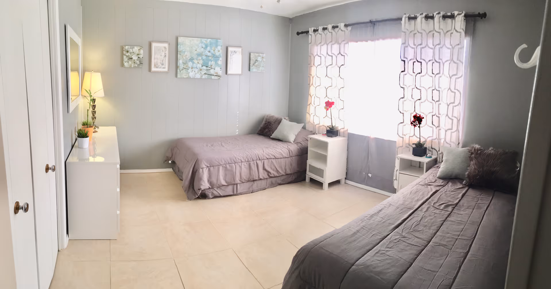 A bright bedroom with two single beds covered in gray bedding, each with pillows and a small nightstand beside them. The room has light-colored tiled floors, gray walls, a large window with patterned curtains letting in natural light, a white dresser with a lamp and small plants, and floral artwork hanging on the wall above one bed.