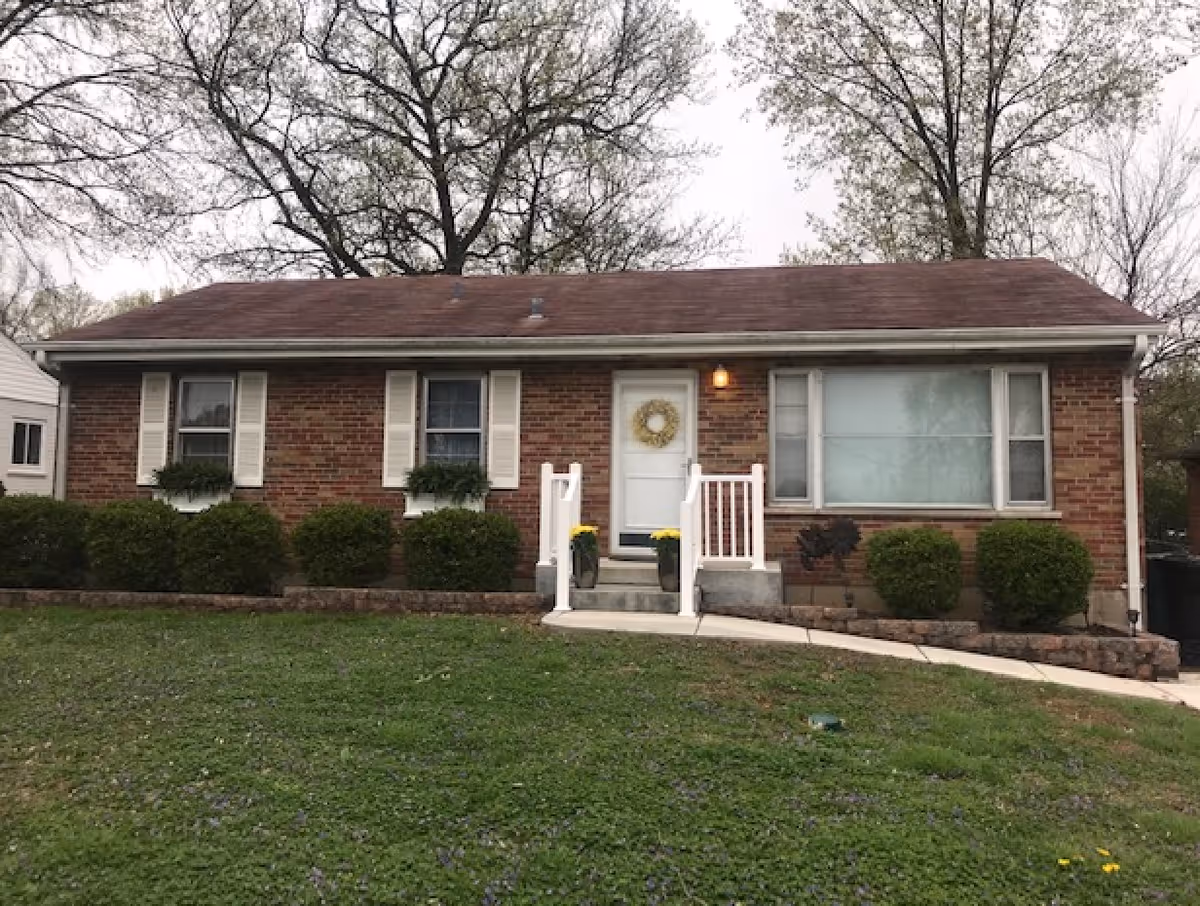 Single-story brick house with a brown roof, white front door with a wreath, two windows with white shutters, and a large window with blinds. There are bushes along the front of the house and a concrete walkway leading to the front steps with white railings. Trees without full leaves are visible in the background.