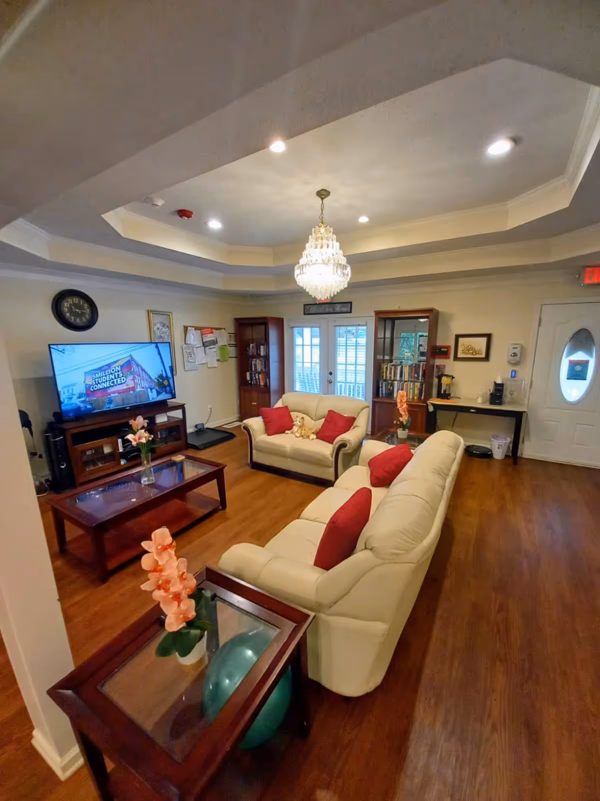 Cozy senior living facility lounge with cream sofas, red pillows, coffee tables, a TV, bookshelves and a chandelier.