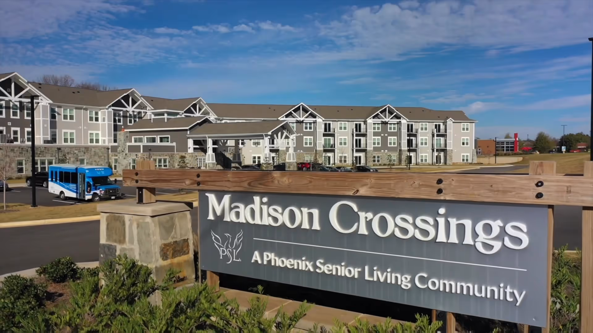 Exterior view of Madison Crossings, a senior living community building with a large sign in the foreground that reads 'Madison Crossings, A Phoenix Senior Living Community'. The building is multi-story with a mix of stone and siding, and a blue shuttle bus is parked on the driveway.