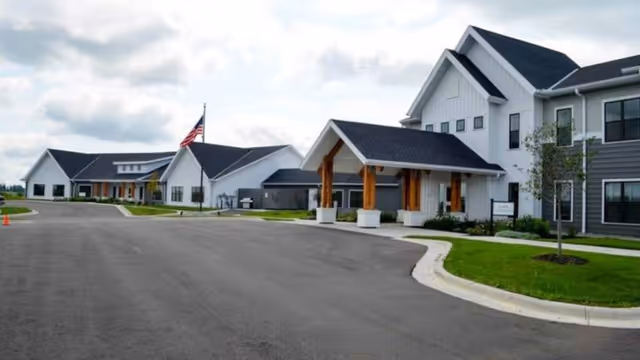 Front exterior of a senior living facility with a wide paved driveway, an American flag, and connected white and gray buildings with covered entrances.