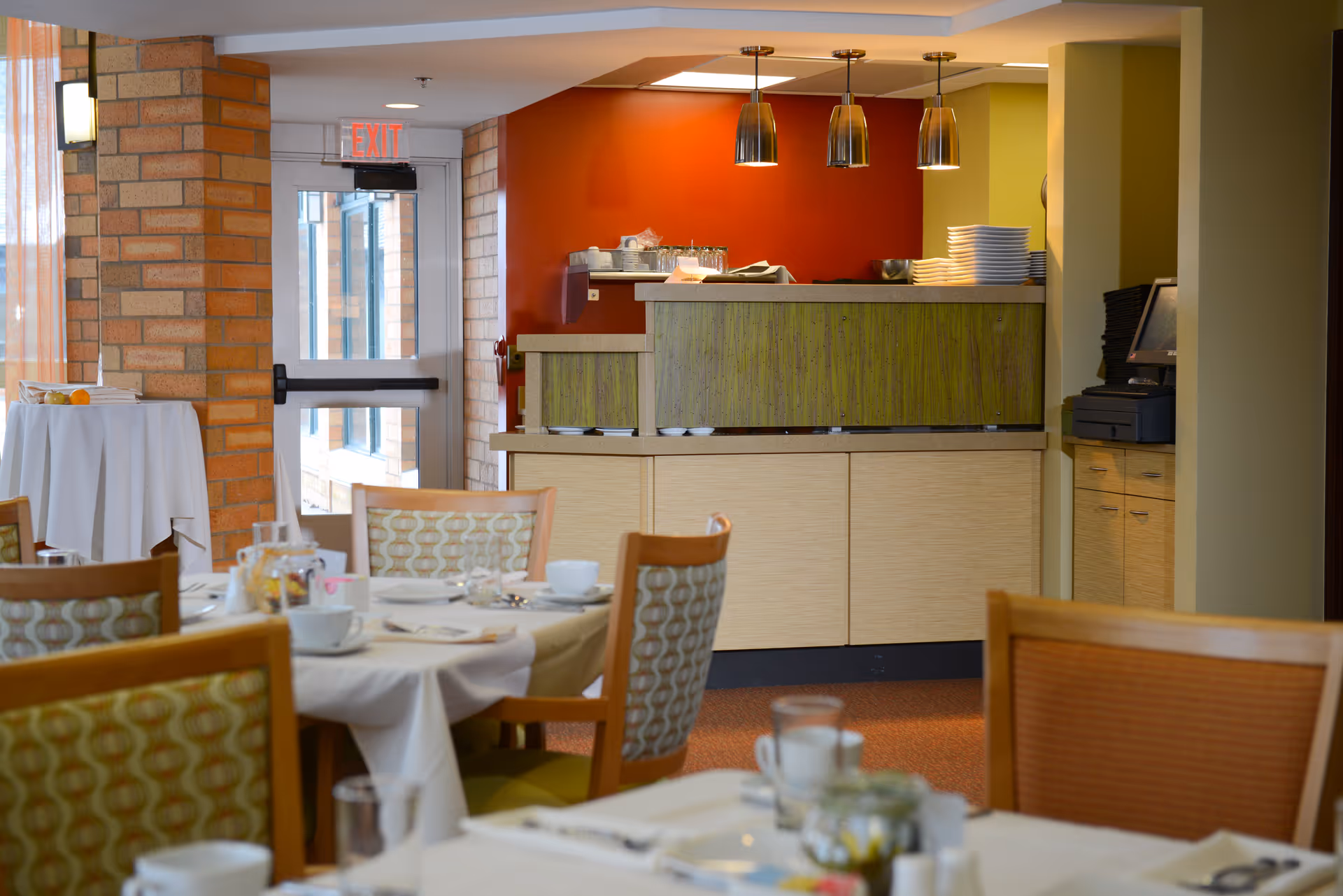A dining area in The Summit at Brighton Independent Living with tables set for a meal, chairs with patterned upholstery, a serving counter with stacked plates, and pendant lights hanging from the ceiling. There is a brick wall and an exit door in the background.