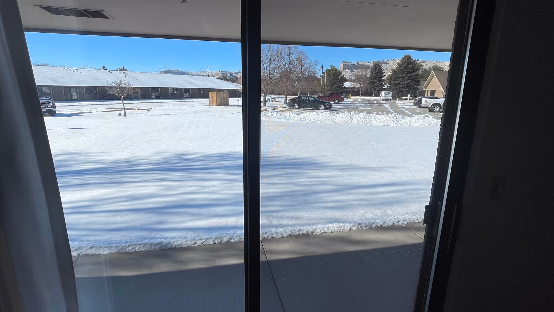 View through a glass sliding door showing a snow-covered lawn, a parking lot with several cars, leafless trees, and buildings in the background under a clear blue sky.