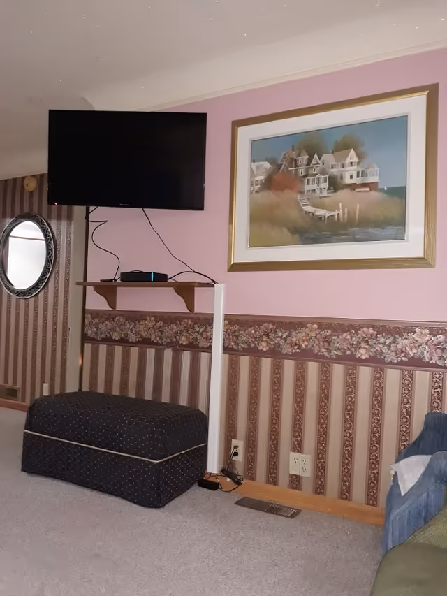 A cozy living room area with a wall-mounted flat-screen TV above a small wooden shelf holding electronic devices. The walls are decorated with floral and striped wallpaper, and a framed painting of houses by the water hangs on the wall. Below the TV is a dark upholstered ottoman, and part of a blue armchair is visible on the right side.