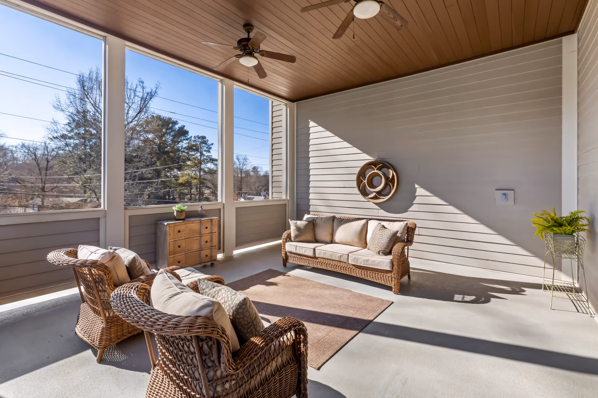 A bright, covered outdoor patio area with wicker furniture including a sofa and two armchairs with cushions, a small wooden chest with a plant on top, a ceiling fan, and a decorative wall piece. The patio has large screened windows showing trees and a clear sky outside.