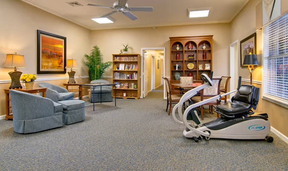 A bright communal living room with upholstered chairs, bookshelves, a dining table, and a recumbent exercise machine by a window.
