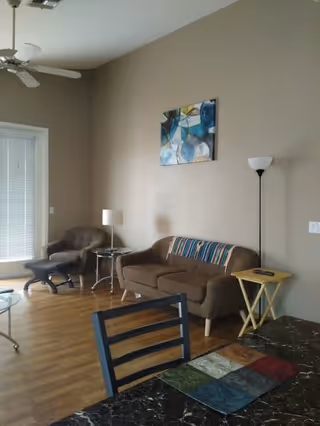 A living room with a brown sofa adorned with a colorful striped throw, a matching armchair with an ottoman, a glass coffee table, a floor lamp, and a side table with a remote control. There is a ceiling fan above and a large window with blinds letting in natural light. A colorful abstract painting hangs on the beige wall.