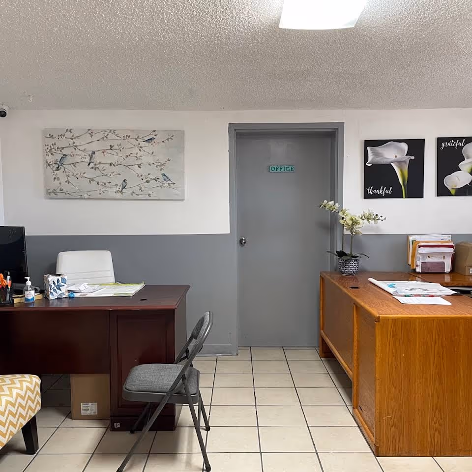 An office area with two desks, one dark wood and one light wood, each with office supplies and paperwork. A gray folding chair is placed in front of the dark wood desk. The wall behind has a gray door labeled 'OFFICE' and three framed artworks featuring flowers and birds. The floor is tiled, and the ceiling has a textured finish with a fluorescent light.