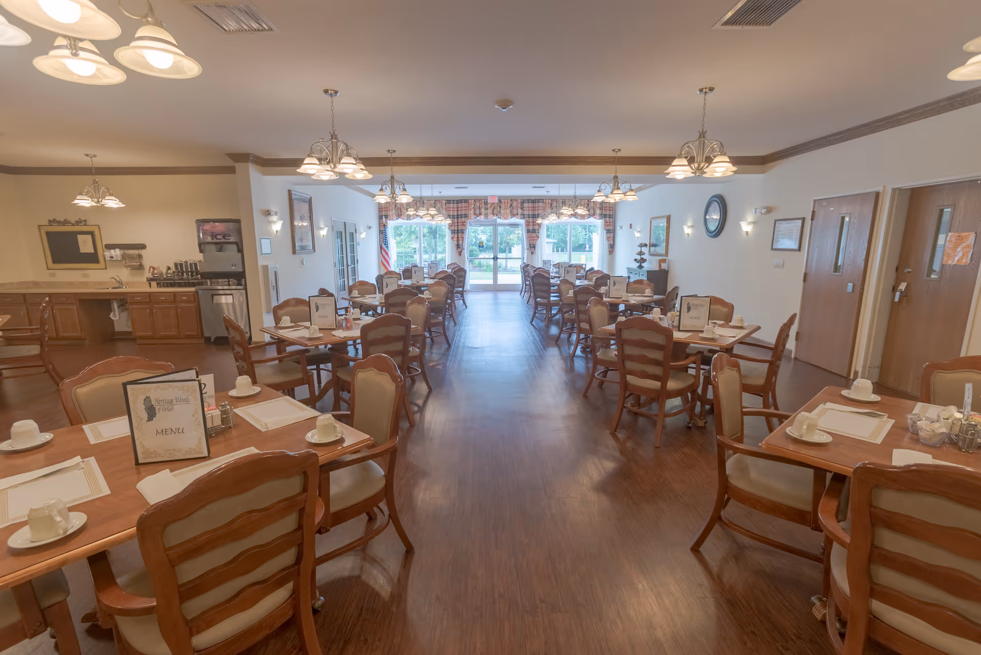 A spacious dining room in a senior living facility with multiple wooden tables and chairs arranged neatly. Each table is set with placemats, cups, and menus. The room has hardwood flooring, soft lighting from ceiling fixtures, and large windows at the far end letting in natural light. There is a small kitchen or service area visible on the left side with cabinets and an ice machine.