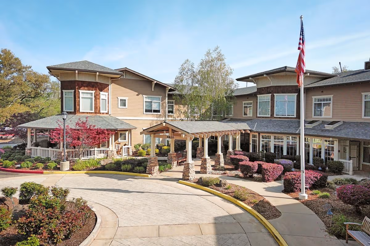 Front exterior view of Sunrise of Fair Oaks senior living facility showing a two-story building with a covered entrance, landscaped garden with bushes and trees, a flagpole with an American flag, and a circular driveway.