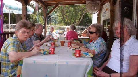 A group of elderly people sitting around a long table covered with a white tablecloth, eating and drinking outdoors under a wooden pergola. Trees and a fence are visible in the background.