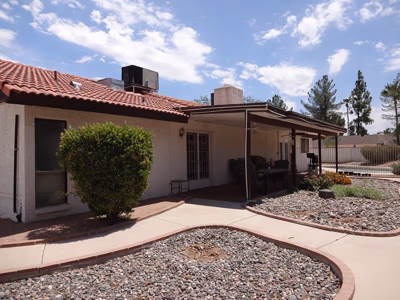 Outdoor view of a single-story building with a red tile roof and a covered patio area. The patio has outdoor furniture and is surrounded by a landscaped area with rocks, a small bush, and some flowers. Trees and a clear sky with clouds are visible in the background.