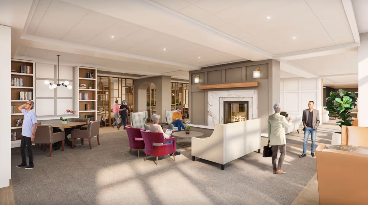 A spacious, well-lit senior living facility common area with comfortable seating including purple and beige armchairs around a coffee table in front of a modern fireplace. Several seniors are engaged in various activities such as reading and conversing. There are bookshelves, a table with chairs, and large windows letting in natural light. The decor is warm and inviting with neutral tones and wood accents.