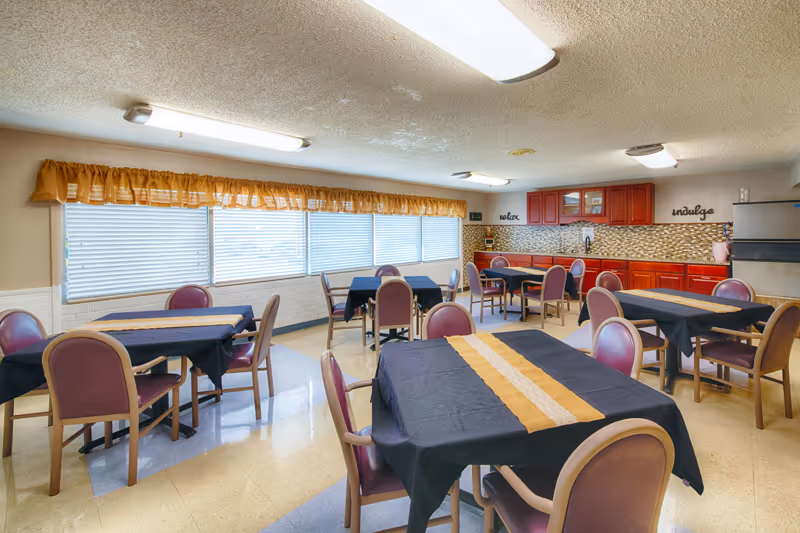 Communal dining room with multiple tables covered in black tablecloths and yellow runners, purple chairs, long windows with a yellow valance, and a kitchenette with red cabinets.