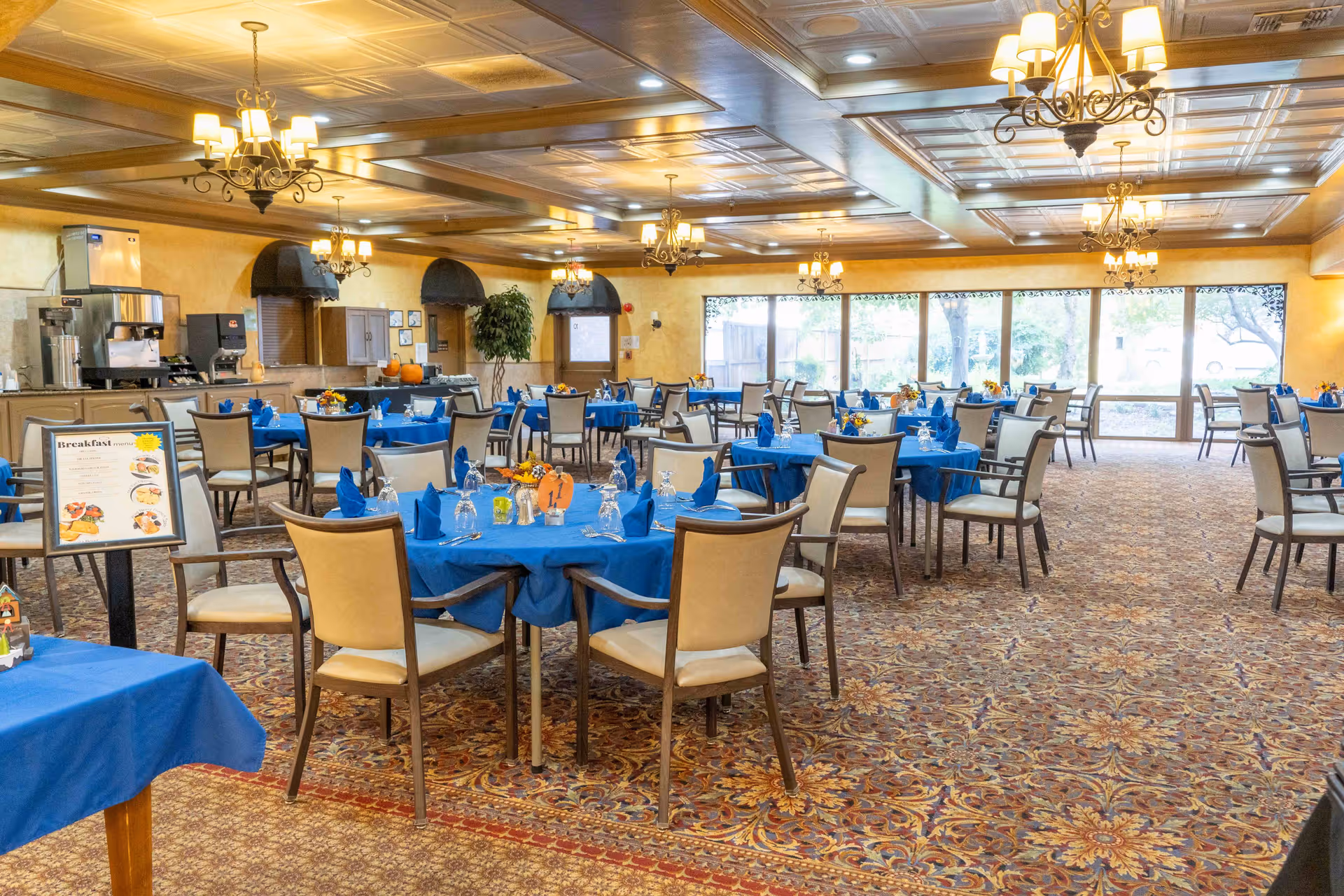 A spacious dining room with multiple round tables covered in blue tablecloths and set with blue napkins, glasses, and silverware. The room features patterned carpet, large windows letting in natural light, chandeliers hanging from a coffered ceiling, and a beverage station along one wall.