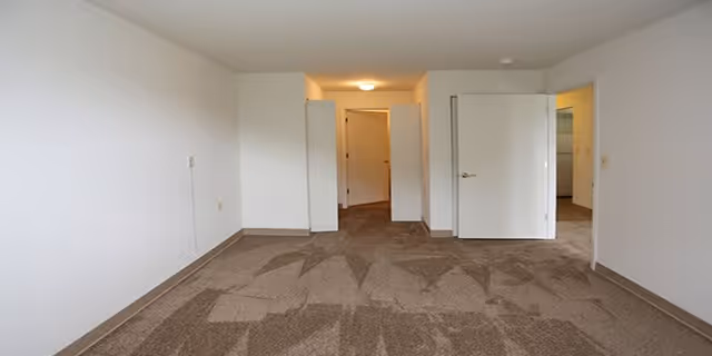 Empty room with beige carpet and white walls, two open white doors leading to other rooms, and a ceiling light fixture in the hallway.