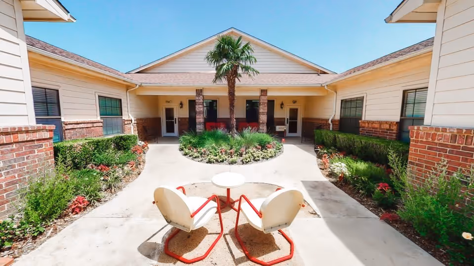 Outdoor courtyard area at Rockbrook Assisted Living and Memory Care featuring a circular garden with a palm tree in the center, surrounded by a concrete walkway. Two white chairs with red frames and a small round table are positioned in the foreground. The courtyard is bordered by a single-story building with beige siding, brick accents, and several windows under a clear blue sky.