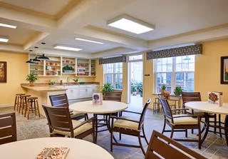 A bright dining room with round tables and wooden chairs arranged neatly. The room has large windows with patterned valances, allowing natural light to fill the space. There is a counter area with bar stools and open shelves displaying plants and decorative items. The walls are painted a warm yellow, creating a welcoming atmosphere.