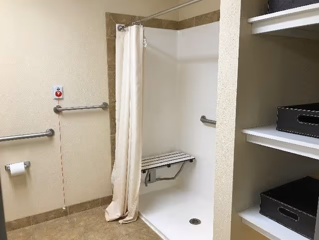 Accessible bathroom featuring a roll-in shower with a fold-down bench and grab bars, a shower curtain, toilet paper holder, emergency pull cord, and open shelving.