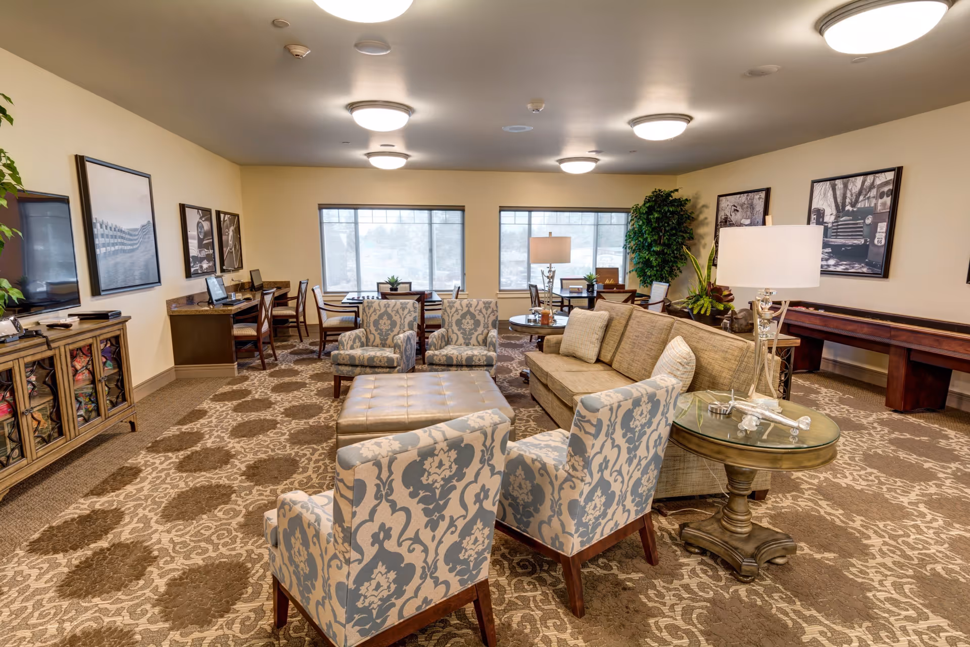 A spacious and well-lit living room area in an assisted living facility featuring patterned armchairs, a beige sofa with pillows, a large ottoman, a round glass side table with a lamp and decorative airplane, and several tables and chairs near windows with blinds. The room has a patterned carpet, framed black and white photographs on the walls, and a large green plant in the corner.