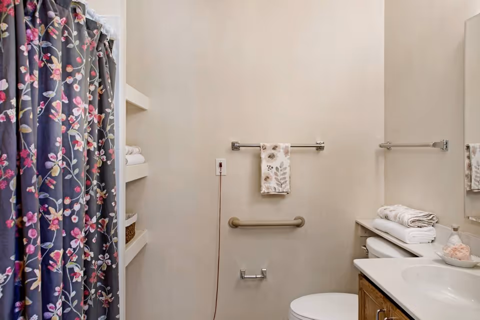 A bathroom with a floral shower curtain on the left, built-in shelves with towels and a basket, a towel rack with a floral hand towel, a grab bar, a toilet, and a countertop with folded towels and a soap dispenser.