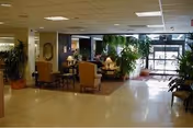 Interior view of a senior living facility lobby or common area with seating arrangements including armchairs and tables, potted plants, lamps, and large windows allowing natural light to enter.