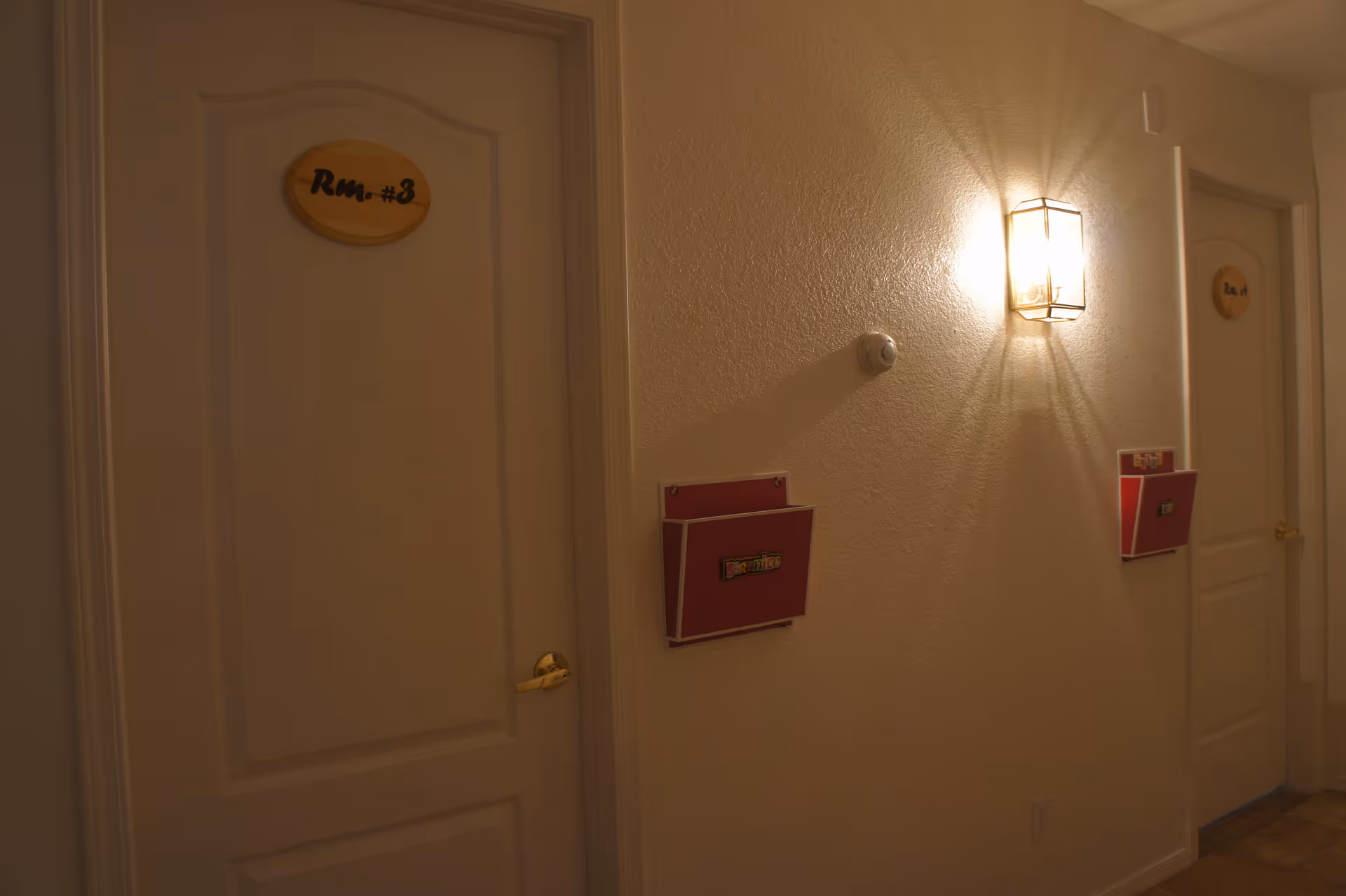 A dimly lit hallway with two closed white doors labeled 'Rm. #3' and 'Rm. #4'. Each door has a golden handle and above each door is a wooden oval sign with the room number. On the wall between the doors is a wall-mounted light fixture casting a warm glow and shadows, along with two red wall-mounted holders.