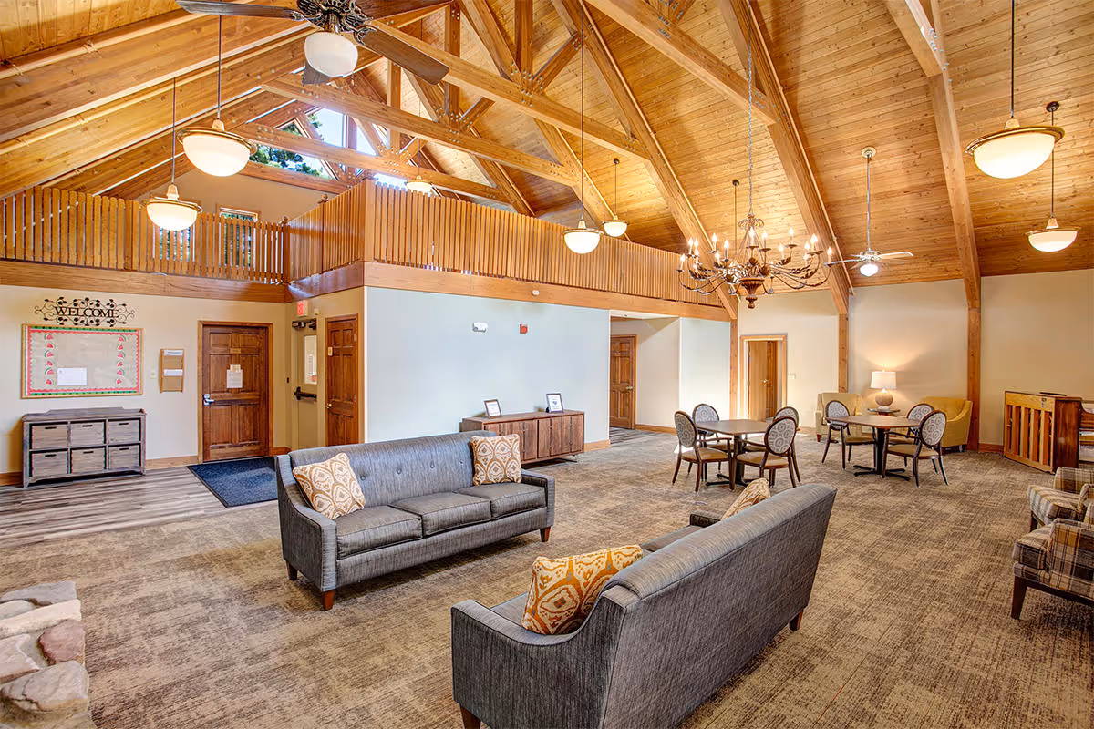 Spacious senior living facility common area with high vaulted wooden ceilings and exposed beams. The room features two gray sofas with patterned pillows, several round tables with chairs, a chandelier, ceiling fans, and warm lighting. There is a wooden railing on the upper level and a welcome board near the entrance door.