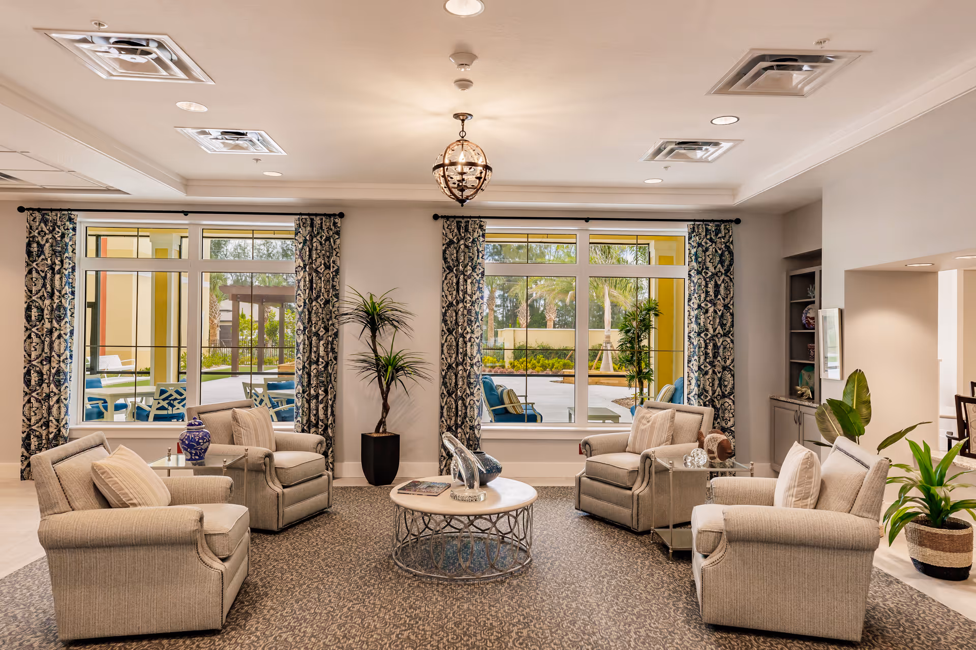 A cozy living room area with six beige armchairs arranged around a circular coffee table. The room features large windows with patterned curtains, allowing natural light to brighten the space. There are several potted plants and decorative items on the table and shelves, creating a welcoming atmosphere. Outside the windows, a patio area with additional seating and greenery is visible.