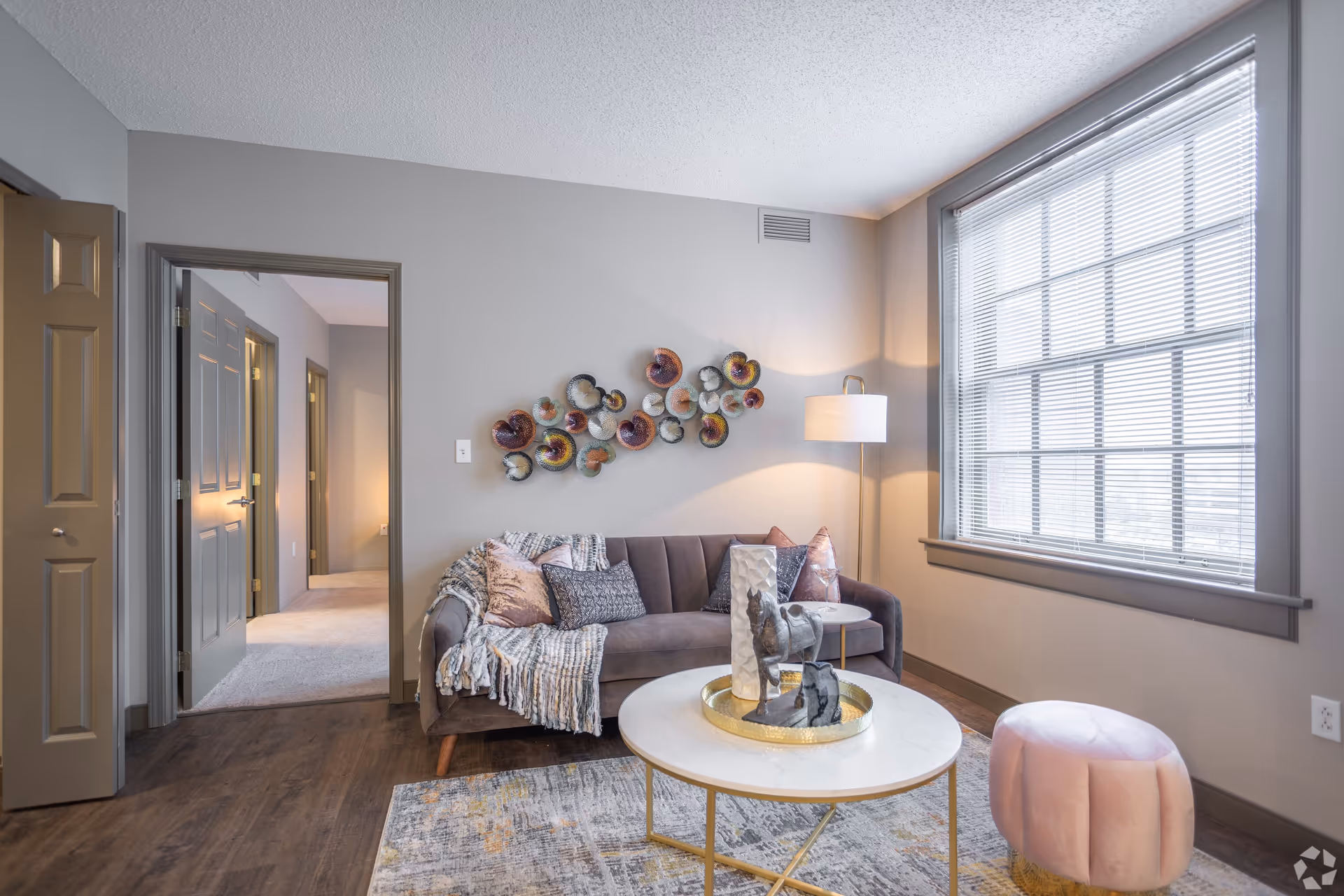 A modern living room with a gray sofa adorned with pillows and a throw blanket, a round white coffee table with decorative items, a pink round ottoman, a floor lamp, a large window with blinds, and a colorful wall art piece. The room has wooden flooring and gray walls with an open doorway leading to another carpeted room.