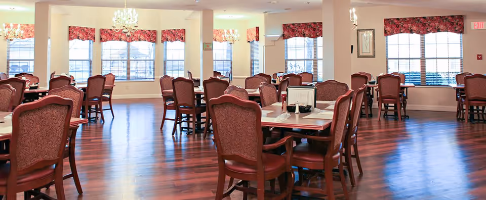 A spacious dining room with multiple wooden tables and upholstered chairs arranged neatly. The room features large windows with floral valances allowing natural light to fill the space, and chandeliers hanging from the ceiling. The floor is polished wood, and the tables are set with menus, napkins, and condiments.