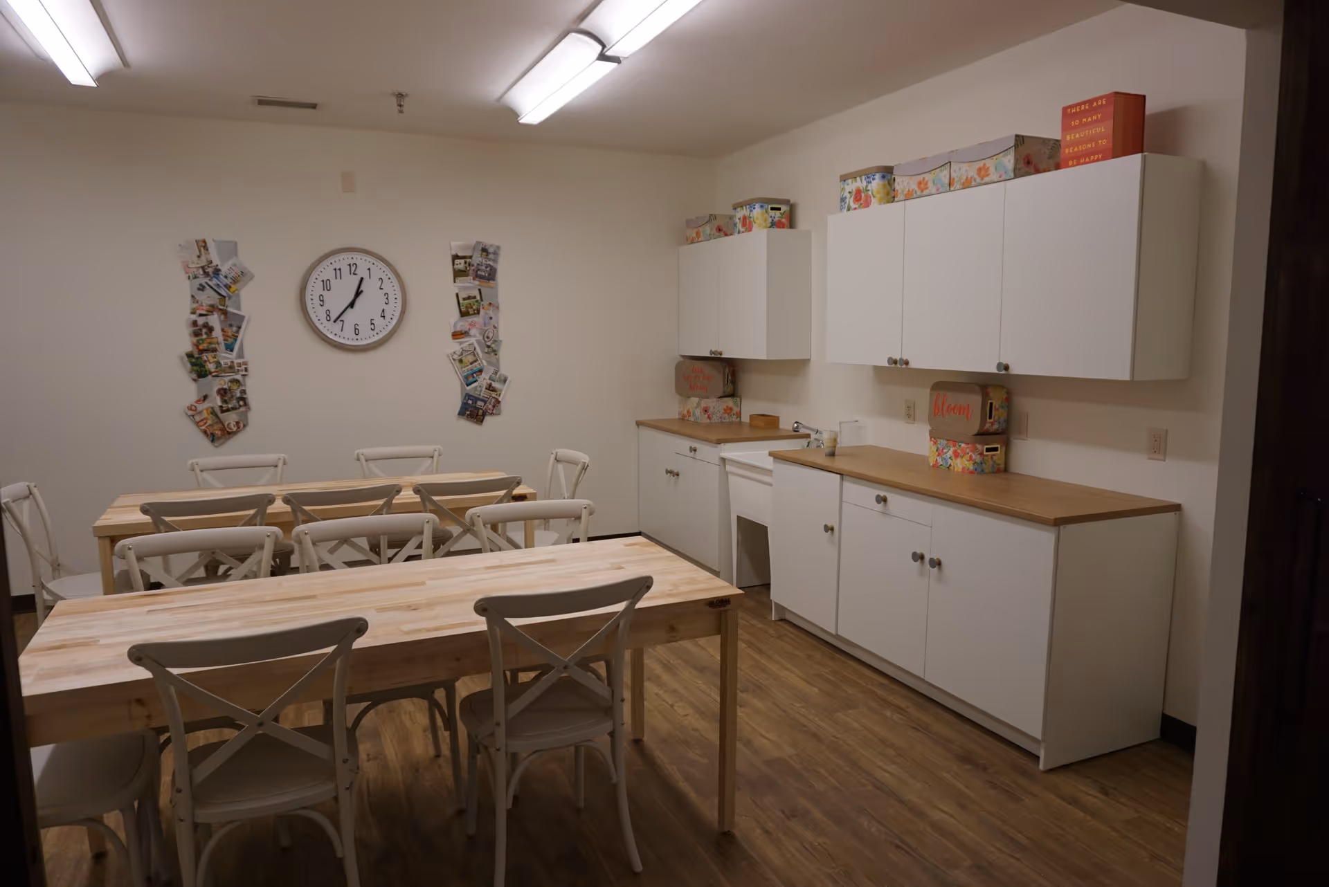 A room with two wooden tables and several white chairs arranged around them. On the right side, there are white cabinets with wooden countertops and decorative boxes on top. A clock is mounted on the wall above the tables, flanked by two vertical displays of postcards or photos. The floor is wooden, and the walls are plain white.