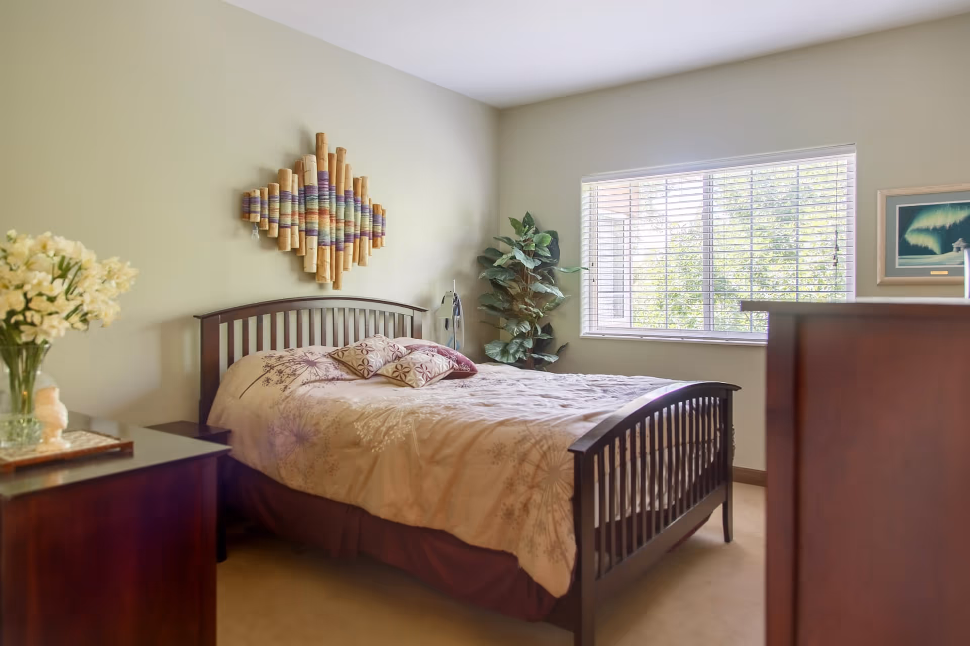 A cozy bedroom with a wooden bed frame and a beige bedspread with floral patterns. There are three decorative pillows on the bed. A wooden nightstand with a vase of flowers is on the left side, and a large window with white blinds lets in natural light. A green potted plant is placed near the window, and a colorful wall decoration hangs above the bed.