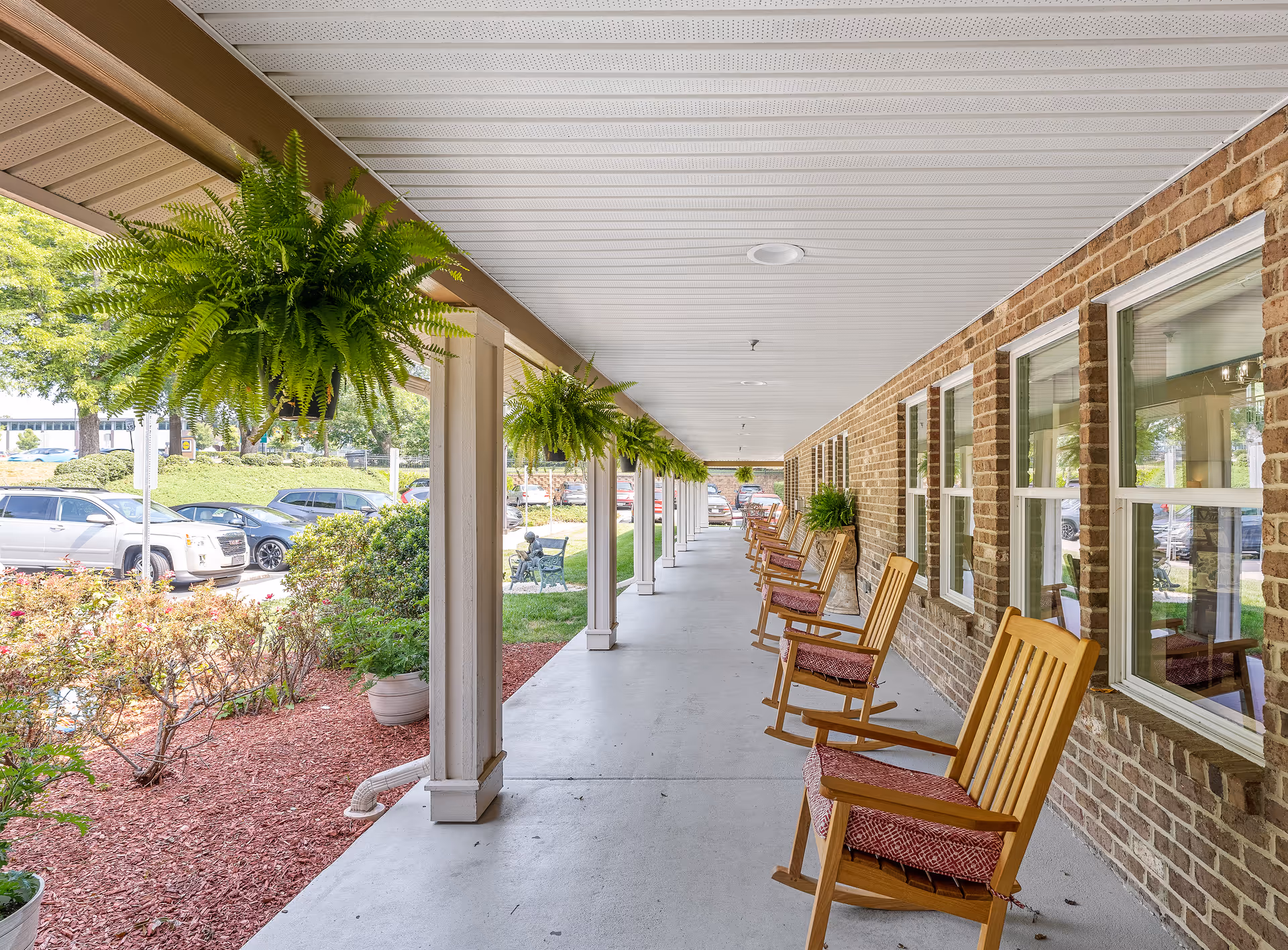 Covered outdoor walkway with hanging green ferns and wooden rocking chairs with cushions lined up against a brick wall with windows. There are cars parked and greenery visible beyond the walkway.