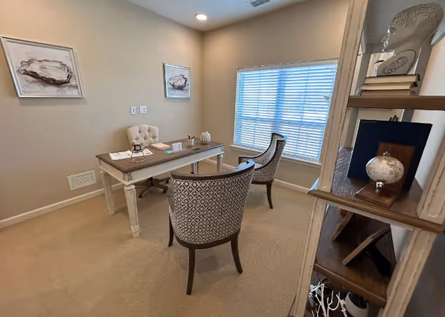 A small furnished office with a desk, three chairs, decorative shelving, and a window with blinds.