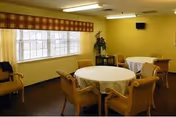 A small dining area with two round tables covered with white tablecloths, surrounded by wooden chairs with cushions. The room has yellow walls, a large window with a plaid valance, a wall-mounted TV, and a floral arrangement in the corner.