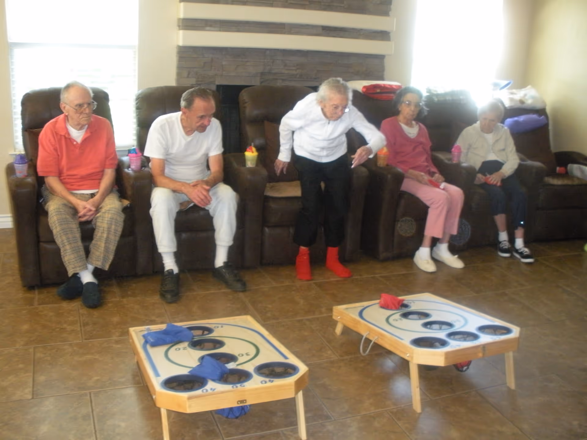 Five elderly people sitting on brown recliner chairs in a living room with tiled floor, playing a bean bag toss game. One elderly woman is standing and throwing a bean bag towards the game boards placed on the floor.