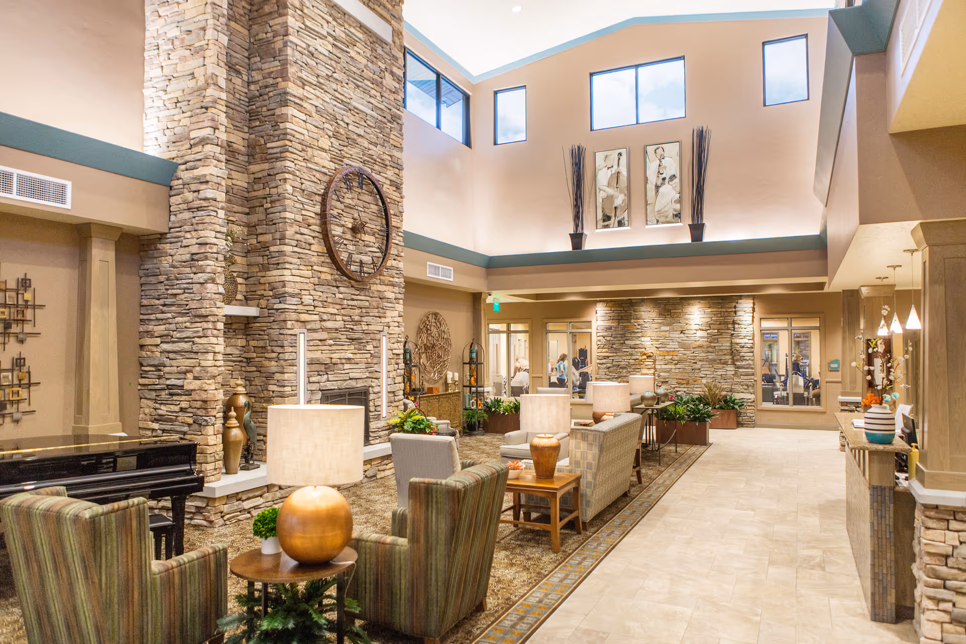 A spacious and well-lit living room area in a senior living facility featuring a tall stone fireplace with a large clock mounted on it. The room has high ceilings with multiple windows, comfortable seating including armchairs and sofas, side tables with lamps, decorative plants, and artwork on the walls. There is a piano on the left side and a reception or service counter on the right.