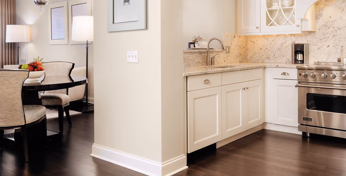 Open kitchen with white cabinets, marble backsplash, stainless steel range, and an adjacent dining table with upholstered chairs and a fruit bowl.