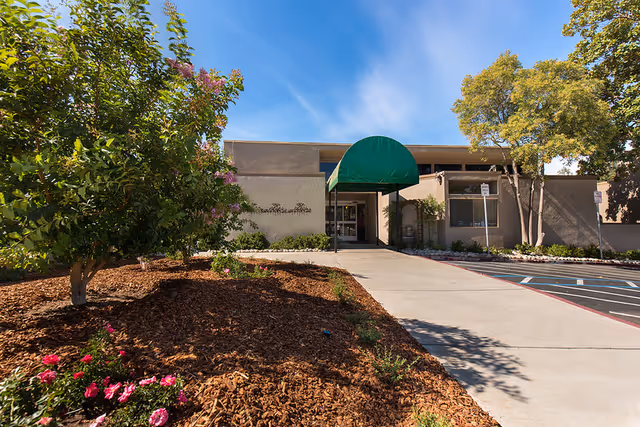 Exterior view of Windsor Manor Rehabilitation Center of Concord showing the entrance with a green awning, surrounded by landscaped garden beds with trees and flowers under a clear blue sky.