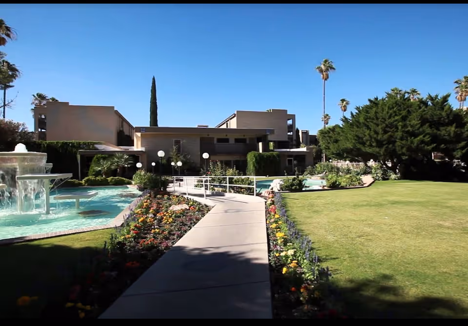 A sunny outdoor scene at Fellowship Square Tucson featuring a paved walkway bordered by colorful flower beds leading to a modern building. There is a water fountain with cascading water on the left side and well-maintained green lawns and trees on the right side under a clear blue sky.