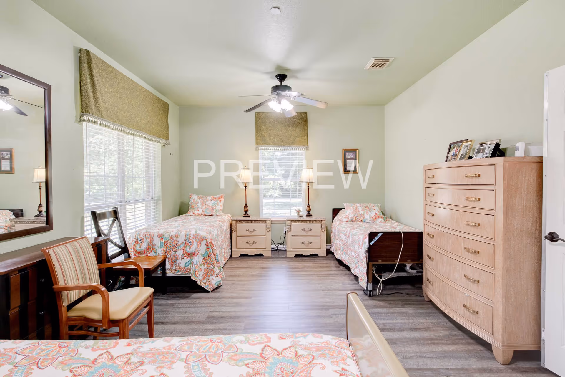 A bright bedroom with three single beds, each covered with colorful patterned bedspreads. The room has light green walls, two windows with beige valances, a ceiling fan with lights, a wooden dresser with framed photos on top, two nightstands between two beds with lamps, and a wooden chair with a cushioned seat near a large mirror on the wall.